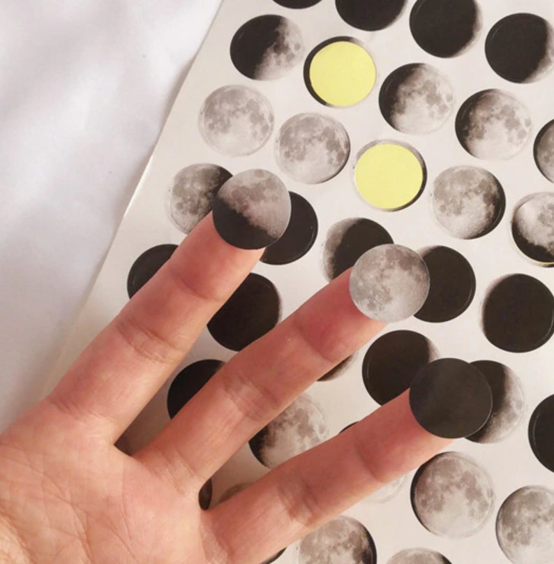 May include: A sheet of moon phase stickers with a variety of moon phases, including a full moon, crescent moon, and new moon. The stickers are black and white and have a matte finish.