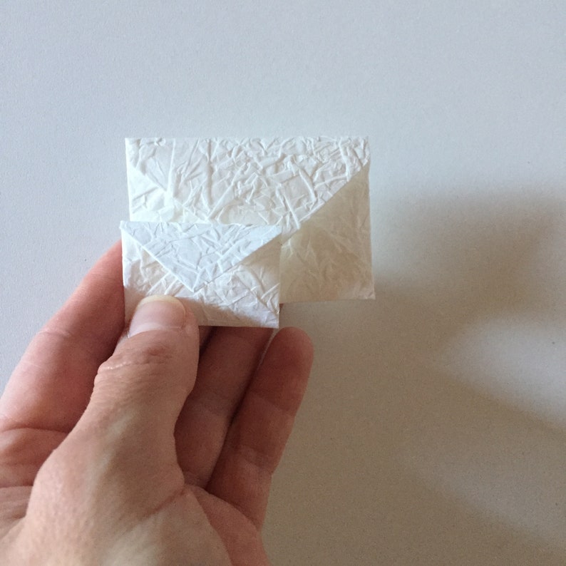 May include: Two white paper envelopes, one nested inside the other, with a textured surface. The envelopes are being held in a hand.