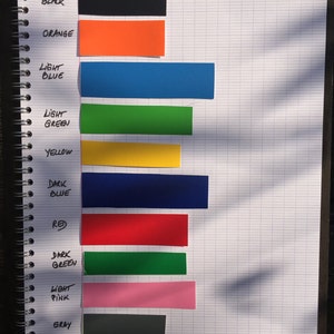 May include: A notebook page with color swatches labeled with the names of the colors: white, black, orange, light blue, light green, yellow, dark blue, red, dark green, light pink, gray, and magenta.