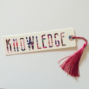Peut inclure: Un marque-page rectangulaire crème avec le mot "KNOWLEDGE" découpé, révélant un motif coloré. Une pampille rose vif est attachée au marque-page avec un petit rivet argenté. Le marque-page est de forme rectangulaire.