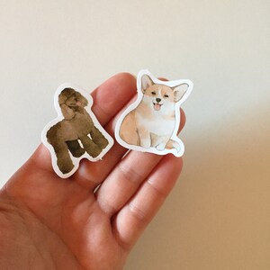 May include: Two dog stickers, one brown poodle and one tan and white corgi, on a hand.