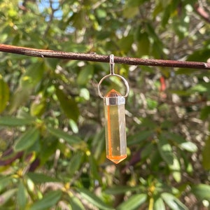 May include: A silver pendant with a clear, orange-tipped crystal. The crystal is a long, hexagonal shape. The pendant hangs from a silver loop, suspended against a backdrop of green foliage.