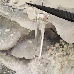 May include: A clear quartz crystal pendant with a silver-colored cap and bail. The crystal is faceted and tapers to a point. The pendant is held by black tweezers with the text "TW-348" printed on it. The background is a light-colored, textured surface.