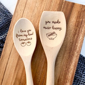 Two twelve inch beech wood utensils are engraved with food related puns. The spoon reads I love you from my head tomatoes and the spatula says you make miso happy with a cute, smiling miso bowl engraved below.
