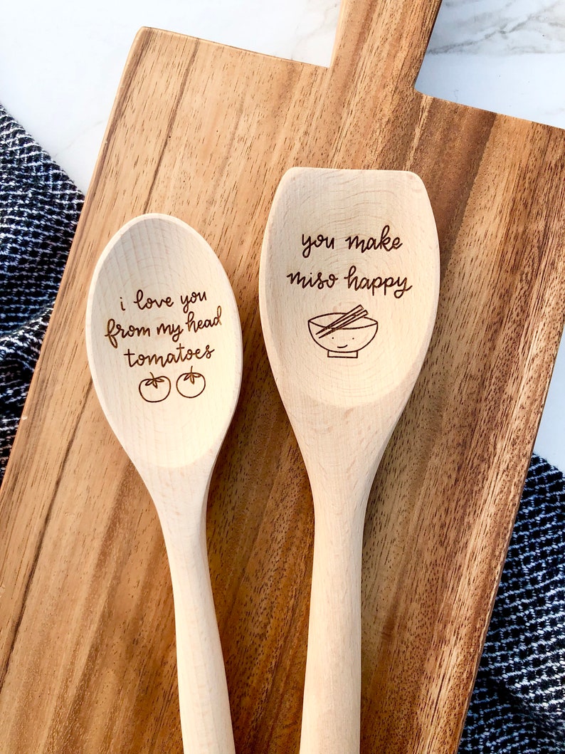 Two twelve inch beech wood utensils are engraved with food related puns. The spoon reads I love you from my head tomatoes and the spatula says you make miso happy with a cute, smiling miso bowl engraved below.