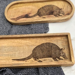May include: Two wooden serving trays with a burned-in design of an armadillo. The larger tray is rectangular and the smaller tray is oval.