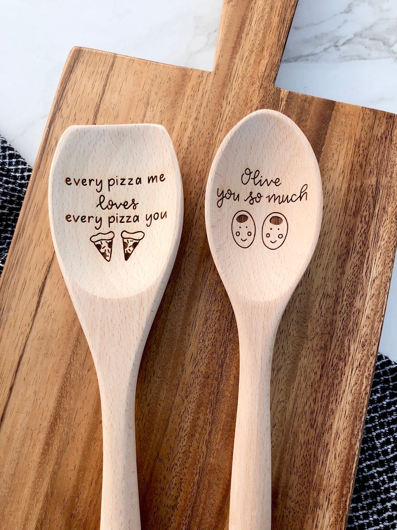 Two twelve inch beech wood utensils are engraved with food related puns. The spoon reads Olive you so much and includes two cute, smiling olives. The wooden spatula says every pizza me loves every pizza you and has two pizza icons engraved below