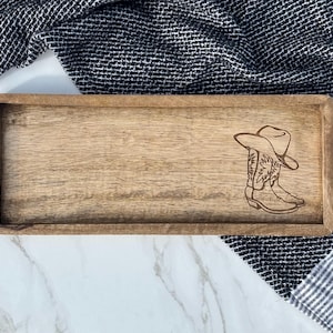 May include: Wooden rectangular tray with a laser-etched design of a cowboy hat and boots. The tray is made of light-colored wood and has a simple, rustic aesthetic. The background includes a black and white patterned cloth.