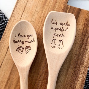 Two twelve inch beech wood utensils are engraved with food related puns. The spoon reads I love you berry much with two strawberries engraved below. The spatula says we make a perfect pear with two smiling pears engraved below the words.