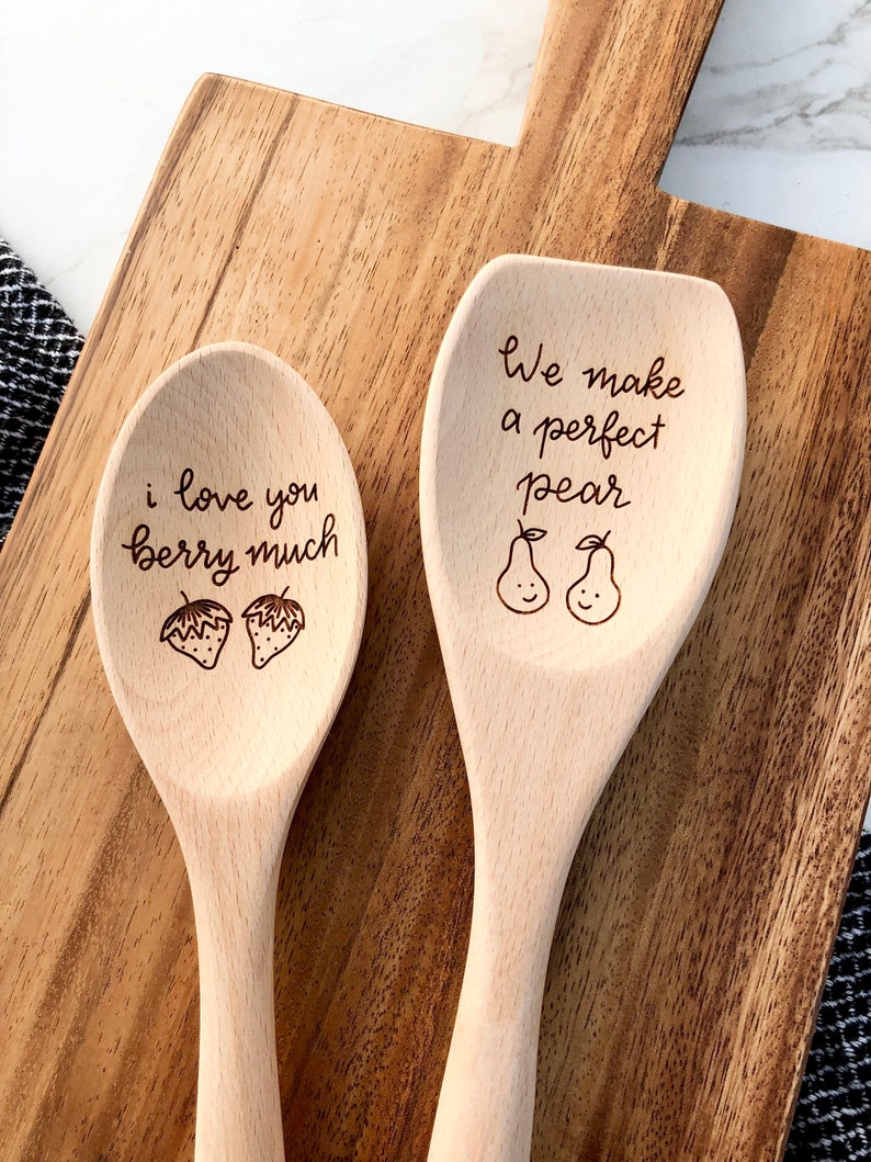 Two twelve inch beech wood utensils are engraved with food related puns. The spoon reads I love you berry much with two strawberries engraved below. The spatula says we make a perfect pear with two smiling pears engraved below the words.