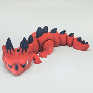 May include: A red and black dragon figurine with a detailed design. The dragon has a long body, with black spikes along its back and head. The dragon is lying down on a white surface.