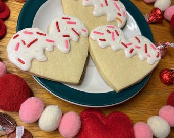 May include: Three heart-shaped sugar cookies decorated with white frosting and red and pink sprinkles on a green plate. The cookies are surrounded by a garland of pink and white felt balls and red felt hearts.