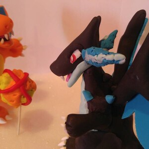 Puede incluir: Dos figuras de Pok&eacute;mon hechas a mano, Charizard y Dragonite, hechas de arcilla polim&eacute;rica. Charizard es naranja con la barriga amarilla y las alas azules. Dragonite es negro con alas azules y una parte inferior azul claro.