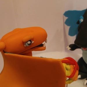 Puede incluir: Primer plano de dos figuras de arcilla de personajes de Pok&eacute;mon. El personaje naranja y azul es Charizard, y el personaje negro y azul es un Pok&eacute;mon parecido a un drag&oacute;n con un ala azul.
