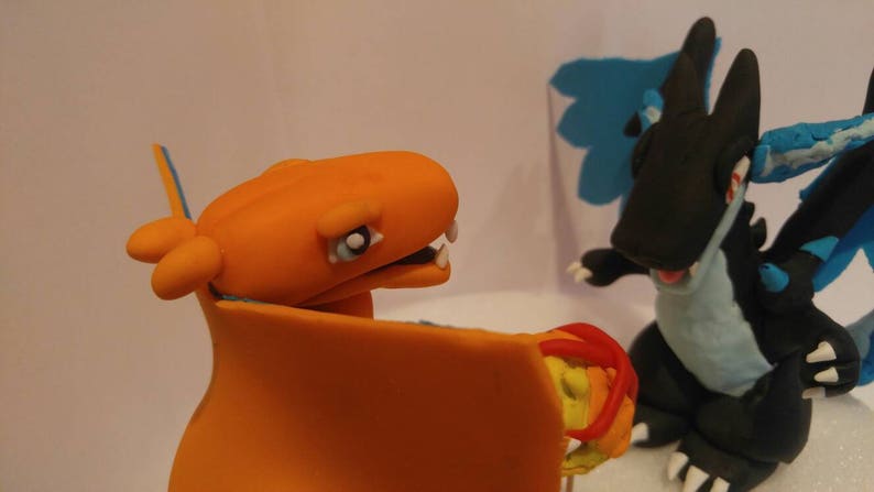 Puede incluir: Primer plano de dos figuras de arcilla de personajes de Pok&eacute;mon. El personaje naranja y azul es Charizard, y el personaje negro y azul es un Pok&eacute;mon parecido a un drag&oacute;n con un ala azul.