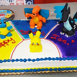Puede incluir: Un pastel cuadrado decorado con glaseado azul, amarillo, morado y blanco. El pastel tiene cuatro secciones, cada una con un personaje de Pokemon diferente en la parte superior. El pastel est&aacute; decorado con las palabras "Happy Birthday Ian!" en glaseado rojo.