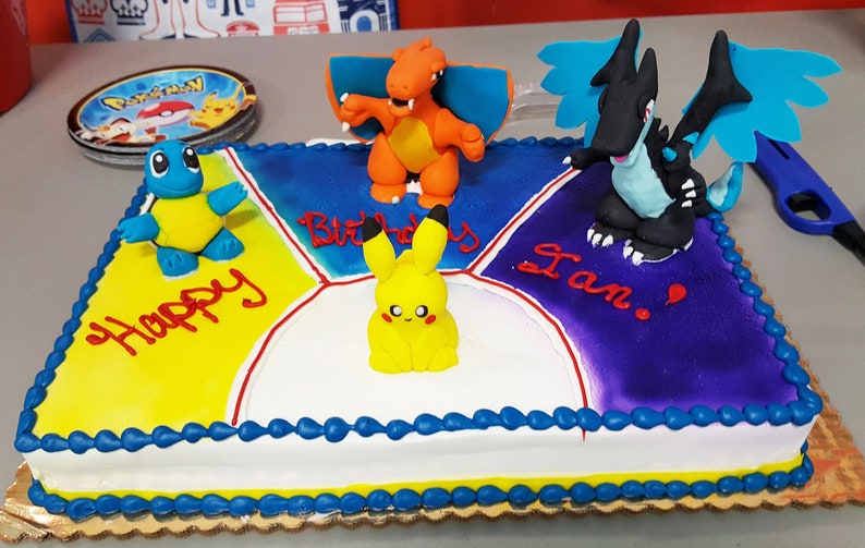 Puede incluir: Un pastel cuadrado decorado con glaseado azul, amarillo, morado y blanco. El pastel tiene cuatro secciones, cada una con un personaje de Pokemon diferente en la parte superior. El pastel est&aacute; decorado con las palabras "Happy Birthday Ian!" en glaseado rojo.