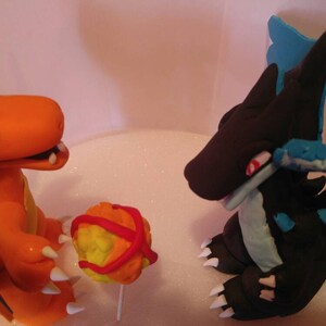 Puede incluir: Dos figuras de Pokemon hechas de fondant. La figura naranja es Charizard, con un ala azul y una Pok&eacute; Ball roja y amarilla. La figura negra es un Dragonite azul y negro con alas azules.