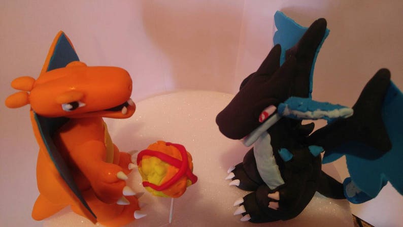 Puede incluir: Dos figuras de Pokemon hechas de fondant. La figura naranja es Charizard, con un ala azul y una Pok&eacute; Ball roja y amarilla. La figura negra es un Dragonite azul y negro con alas azules.