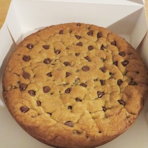 May include: A large, round chocolate chip cookie in a white cardboard box. The cookie is golden brown and has a crispy texture. It is covered in chocolate chips.