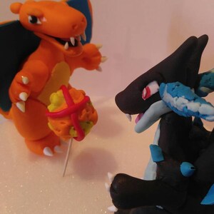 Puede incluir: Dos figuritas hechas a mano de personajes Pok&eacute;mon, Charizard y Rayquaza. Charizard es naranja con detalles amarillos y sostiene una Pok&eacute; Ball roja y amarilla. Rayquaza es negro con detalles azules y tiene ojos rojos.
