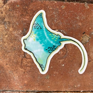 May include: A teal blue and white patterned sticker of a stingray with a yellow outline. The stingray has a long tail and black spots.
