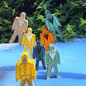 Puede incluir: Un conjunto de coloridos pines con forma de figuras en varios colores, incluyendo amarillo, naranja, verde y beige. Cada pin presenta una cara sonriente y un diseño de traje. Los pines están dispuestos sobre una superficie azul con un fondo de follaje verde.