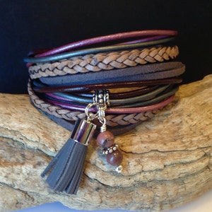 May include: A multi-layered leather bracelet with a braided leather strap and a tassel charm. The bracelet features a variety of colors, including brown, gray, and pink. The tassel is made of gray leather and has a silver bead at the end. The bracelet is accented with a silver charm and two pink stones.
