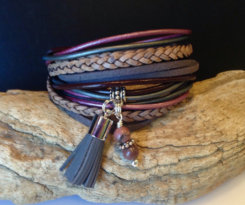 May include: A multi-layered leather bracelet with a braided leather strap and a tassel charm. The bracelet features a variety of colors, including brown, gray, and pink. The tassel is made of gray leather and has a silver bead at the end. The bracelet is accented with a silver charm and two pink stones.