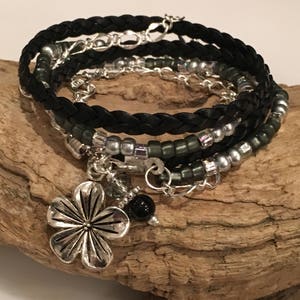 May include: A black braided leather bracelet with silver and green beads and a silver flower charm. The bracelet is layered with a silver chain.