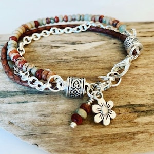 May include: A brown leather bracelet with a silver chain and a silver flower charm. The bracelet is decorated with multi-colored stones.