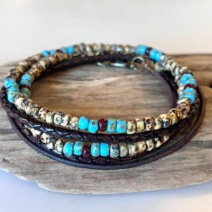 May include: A multi-strand wrap bracelet featuring dark brown braided leather cords interspersed with small, round beads in turquoise blue, deep maroon, and speckled cream and brown patterns, secured with a metallic hook and loop clasp.