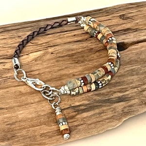 Puede incluir: Una pulsera de múltiples hilos con una correa de cuero trenzado marrón y un cierre. La pulsera tiene dos hilos de pequeñas cuentas rectangulares de colores en tonos marrón, gris y beige. Un pequeño dije cuelga del cierre.