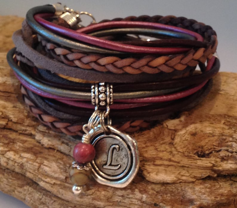 May include: A multi-strand leather bracelet with a braided brown leather strap and a silver charm with the letter "L" on it. The charm is attached to a silver chain with a small red stone bead.