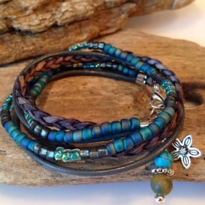 May include: A multi-strand wrap bracelet featuring blue and green beads, braided brown leather, and a gray leather band. A silver flower charm and a dangling bead add detail. The bracelet is displayed on a piece of driftwood.