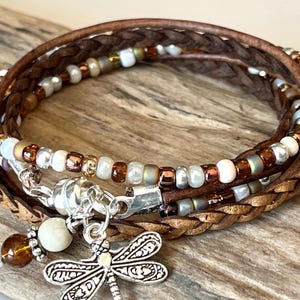 May include: A brown leather wrap bracelet with a dragonfly charm. The bracelet is made with multiple strands of leather and beads in shades of brown, white, and gold.