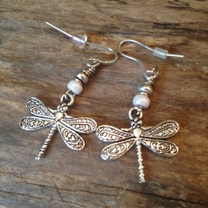 May include: Silver dragonfly earrings with white pearl accents. The earrings have a delicate, intricate design and are perfect for adding a touch of whimsy to any outfit.