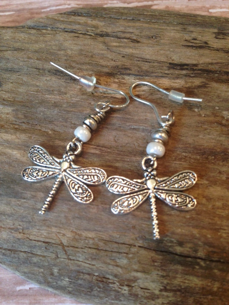 May include: Silver dragonfly earrings with white pearl accents. The earrings have a delicate, intricate design and are perfect for adding a touch of whimsy to any outfit.