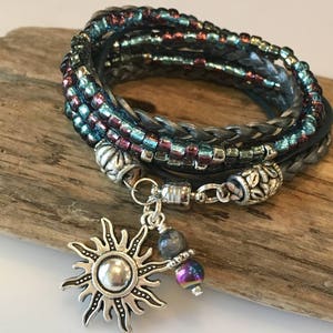 May include: A multi-strand bracelet with blue, silver, and burgundy beads, a braided leather cord, and a silver sun charm with a dark blue and iridescent purple bead.