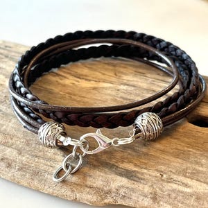May include: Braided leather bracelet in shades of brown and black. The bracelet features multiple strands of leather, with a silver-colored clasp and decorative beads. The bracelet is designed to wrap around the wrist multiple times.