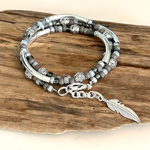 Puede incluir: Pulsera envolvente de múltiples hilos con cuentas grises y plateadas, acentuada con cuentas y tubos plateados decorativos. La pulsera tiene un dije de pluma plateada y un cierre de broche. Se muestra sobre un trozo de madera.