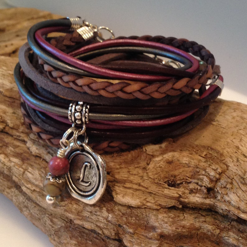 May include: A multi-layered leather bracelet with a braided brown leather strap and a silver charm with the letter "L" and a red stone.