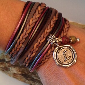 May include: A multi-strand leather bracelet with a braided brown leather strap and a silver charm with the letter "L" on it. The bracelet is adorned with beads and has a silver clasp.