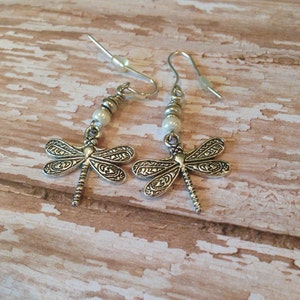 May include: A pair of silver dragonfly earrings with white pearl beads. The earrings have a delicate design and are perfect for adding a touch of whimsy to any outfit.