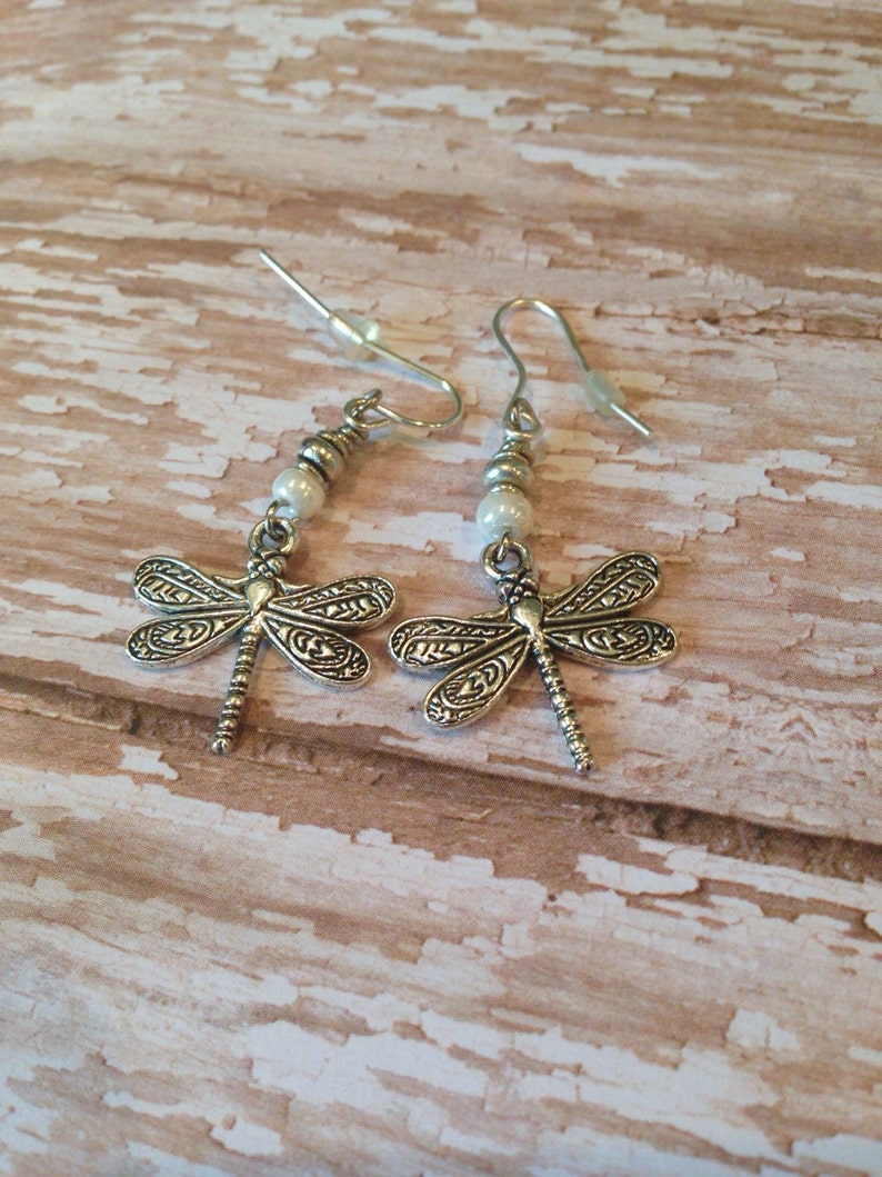 May include: A pair of silver dragonfly earrings with white pearl beads. The earrings have a delicate design and are perfect for adding a touch of whimsy to any outfit.