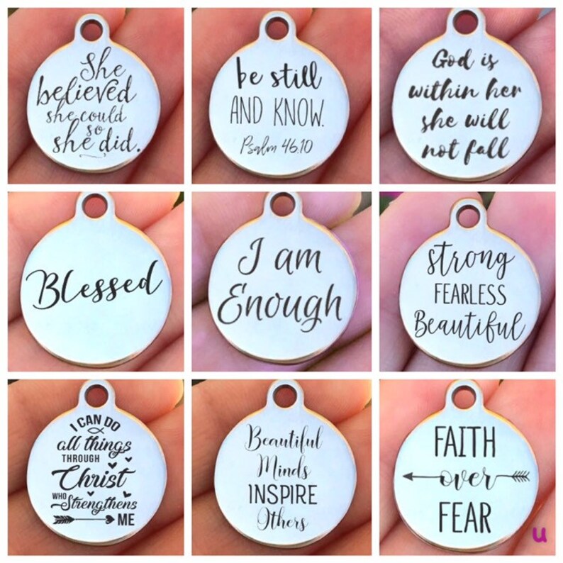 May include: Nine silver charm pendants with inspirational quotes and phrases. The charms are round and have a hole at the top for attaching to a chain or bracelet. The quotes include "She believed she could so she did", "Be still and know", "God is within her she will not fall", "Blessed", "I am Enough", "Strong Fearless Beautiful", "I can do all things through Christ who strengthens me", "Beautiful Minds Inspire Others", and "Faith over Fear".