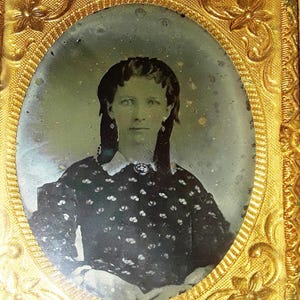 May include: A black and white daguerreotype portrait of a woman with dark hair, wearing a patterned dress. The portrait is framed in a gold-colored frame with ornate details.