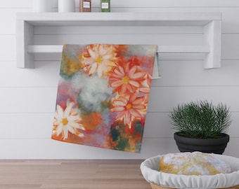 May include: A floral kitchen towel with a watercolor design of orange and white flowers. The towel hangs on a white shelf above a wooden countertop. The background includes a white wall, a small potted plant, and a basket of bread.