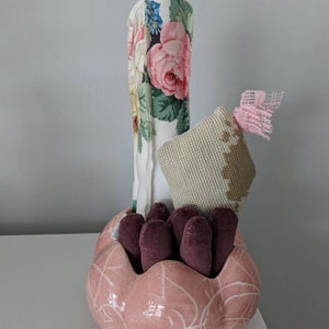 Peut inclure: Composition décorative. Un bol en céramique rose avec un motif veiné blanc contient des objets violets foncés. Un objet cylindrique et long est recouvert d'un tissu à motifs floraux, et un élément en tissu beige avec un nœud rose est également présent.