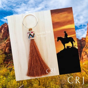 May include: A brown tassel keychain with a beaded charm that says "Apache Trail by K. Morgan". The background is a wooden surface with a sunset scene in the background.